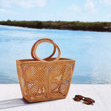 A woven rattan handbag featuring a butterfly pattern rests on a surface near the water's edge, surrounded by green plants and sunglasses. The bag showcases a trapezoid shape with intricate wicker details, complemented by real rattan handles made from Ata grass. This eco-friendly, fair-trade accessory exudes natural elegance and modern style.