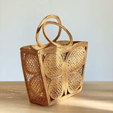 A woven rattan handbag featuring a butterfly pattern rests on a wooden table, showcasing its trapezoid shape and circular, woven handle made of rattan. The bag's unique design and eco-friendly material make it a stylish accessory for everyday use.