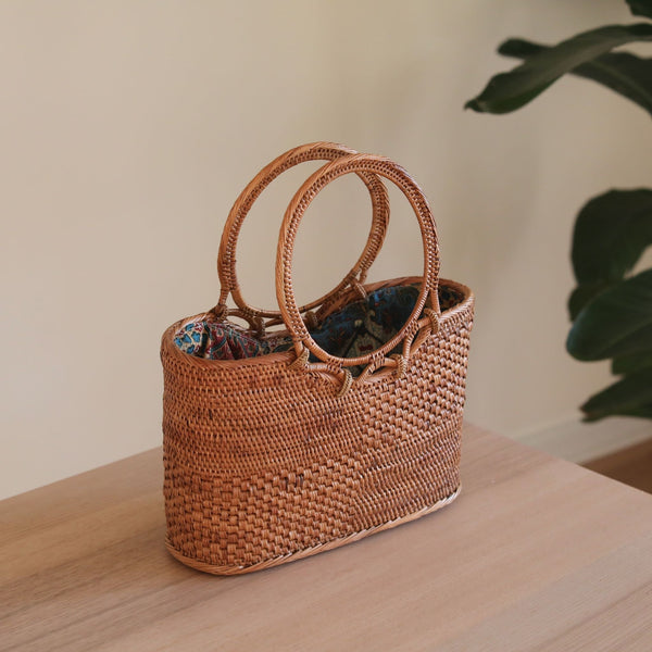 A woven rattan handbag, Bali Rattan Handbag - Kelsey, rests on a surface. The bag is made from natural materials like ata grass for the circular handles, giving it a textured appearance. This stylish yet functional accessory features a spacious interior and a batik lining drawstring pouch.