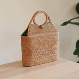 A cylindrical-shaped rattan handbag resting on a neutral background, showcasing its natural materials and woven texture adorned with geometric patterns. The bag features a straightforward style, highlighting its unique design and craftsmanship.