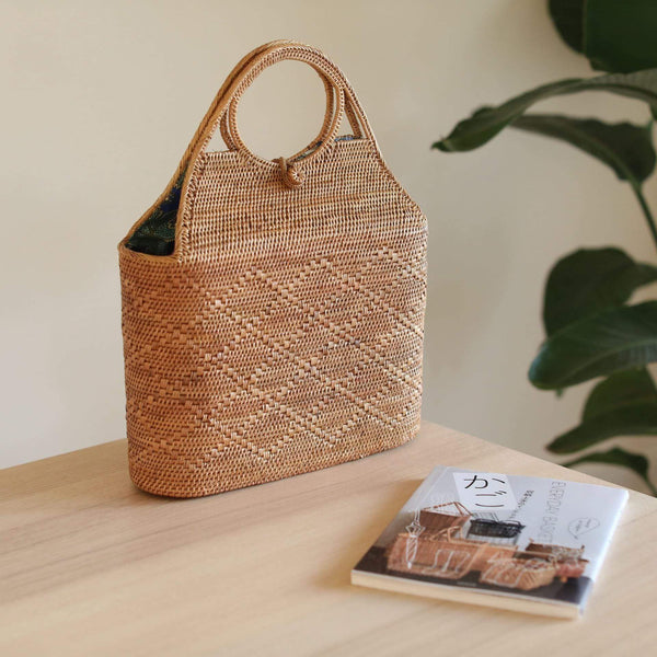 A cylindrical-shaped Bali Rattan handbag rests on a flat surface, surrounded by a book and a small plant, evoking a casual homey setting. The handbag features a woven texture made from natural materials, with a unique cylindrical shape and a beautiful Batik lining visible inside.