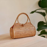 A woven rattan handbag, cylindrical in shape, sits on a wooden surface amidst lush greenery. The bag's natural texture and earthy tones are highlighted by the surrounding plants. Made from rattan and bamboo, it features a sturdy handle for carrying. The image conveys simplicity and elegance.