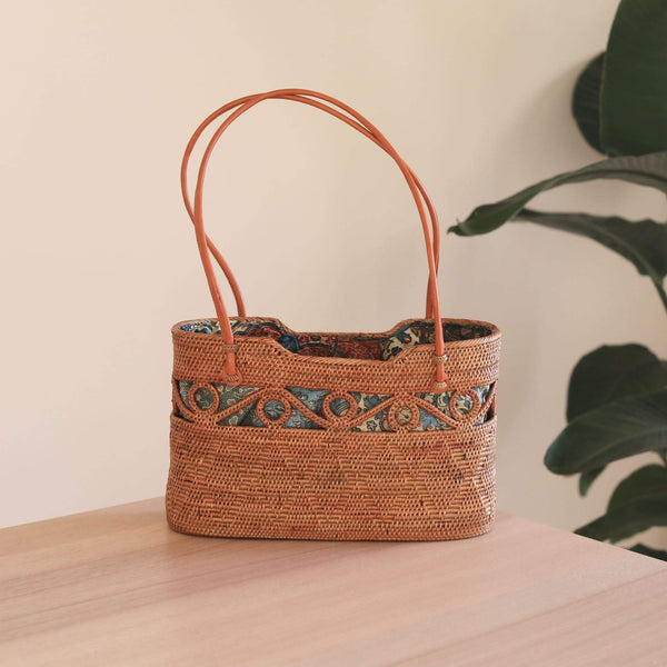 A beautifully crafted Bali Rattan Handbag, the Roma model, sits on a wooden surface, showcasing its woven texture in shades of brown and beige. The oval-shaped handbag features a prominent leather handle on one side, adding a touch of elegance to its design. Made from eco-friendly materials and handmade by skilled artisans in Bali, this unique bag boasts a Batik lining for added uniqueness.