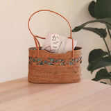 A woven handbag, oval-shaped when viewed from the top, rests on a surface amidst an indoor setting. The bag's rattan material and real leather handle are visible, with a batik lining inside. The overall mood is calm and inviting.