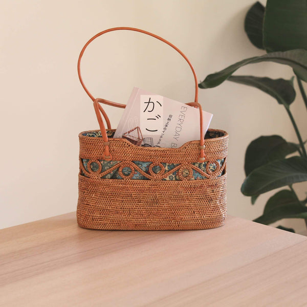 A woven handbag, oval-shaped when viewed from the top, rests on a surface amidst an indoor setting. The bag's rattan material and real leather handle are visible, with a batik lining inside. The overall mood is calm and inviting.