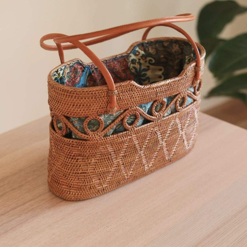 A handbag with a brown handle rests on a wooden surface, showcasing its circular shape and decorative pattern. The bag's interior is lined with vibrant batik fabric, contrasting with the neutral woven material. The simple yet stylish design highlights the handbag as the main focus.