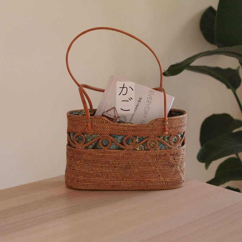 A woven basket with a handle on top, placed on a surface, made from natural fibers with a circular shape when viewed from the top. The background features a simple indoor setting with a plant visible in the corner, suggesting a home environment.