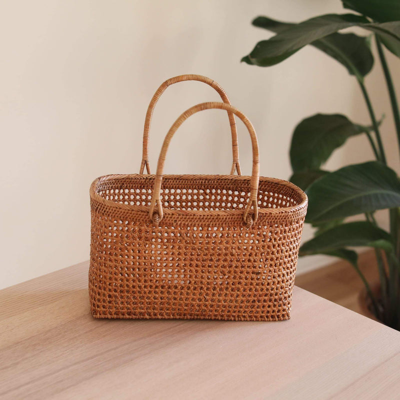 A woven rattan handbag rests on a table with natural textures, showcasing its classic design and solid handle. The bag features visible weaving patterns and a light color scheme, crafted from natural materials. A simple indoor setting with a plant in the corner complements the product's natural aesthetic.