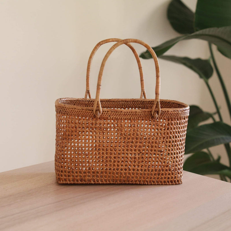 A woven rattan handbag sits on a flat surface, its natural brown color and visible weaving patterns taking center stage. The bag's unique texture and design are showcased against a simple background featuring a plant and white wall.