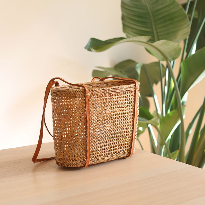 A rectangular woven rattan bag rests on a wooden surface, its rounded corners and handle at one end visible. The natural material features an intricate weaving pattern, with a plant in the background adding greenery to the scene.