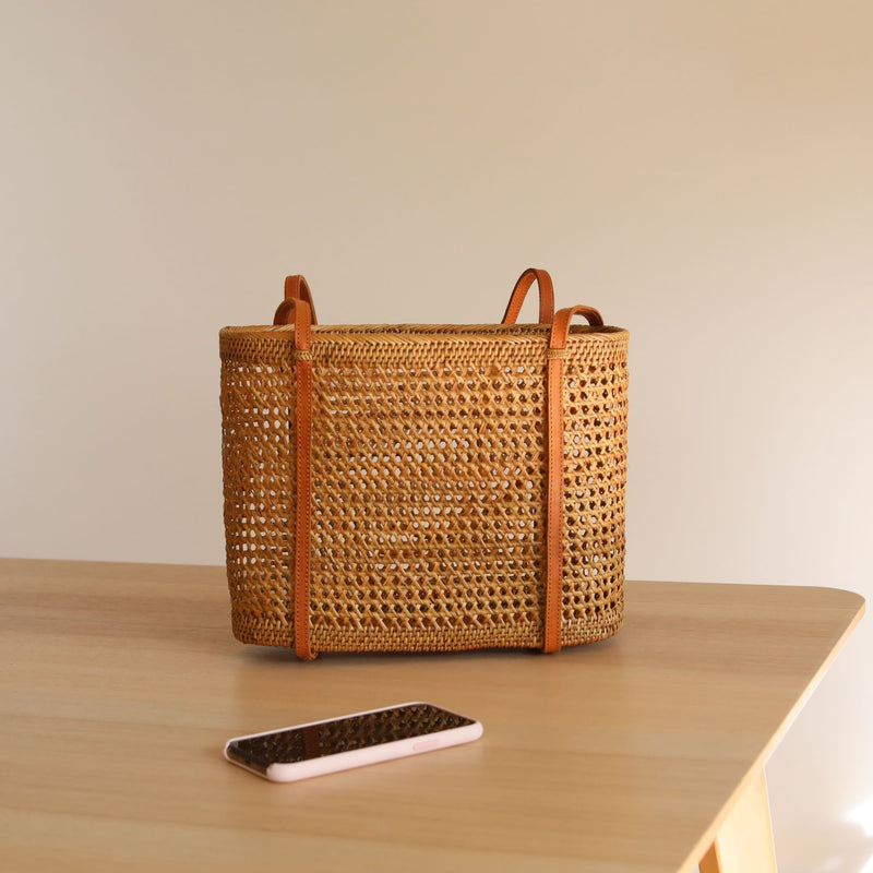 A light brown wicker tote bag rests on a wooden surface, showcasing its textured weave pattern and two leather handles. The spacious interior is empty, highlighting the bag's stylish design.