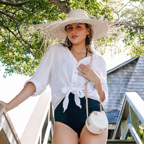 A woman stands outdoors on a sunny day, wearing a white blouse and black shorts, paired with a wide-brimmed straw hat matching her bag. The Bali Mini Rattan Crossbody Bag has a rattan texture and a bow at the front, resting against her hip. The natural setting of trees and a wooden structure creates a casual and leisurely mood.