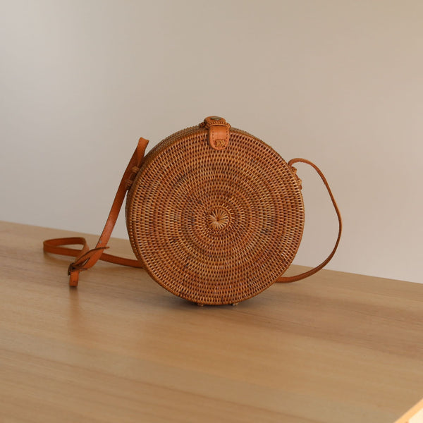 A woven rattan crossbody bag in brown, placed on a wooden surface, featuring an adjustable leather strap, batik lining with a circular pattern, and a secure snap closure. This eco-friendly bag is made from natural rattan and handwoven by artisans in Bali.