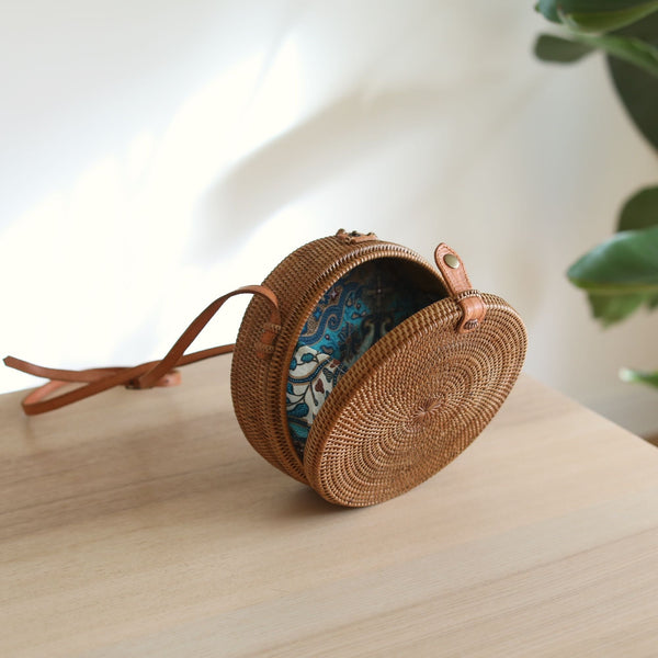 A woven rattan crossbody bag with a round shape sits on a wooden surface, surrounded by a plant. The adjustable leather strap is visible, and the batik lining can be seen through the opening. Handmade from natural rattan materials, this eco-friendly accessory features real leather straps and a secure snap closure.