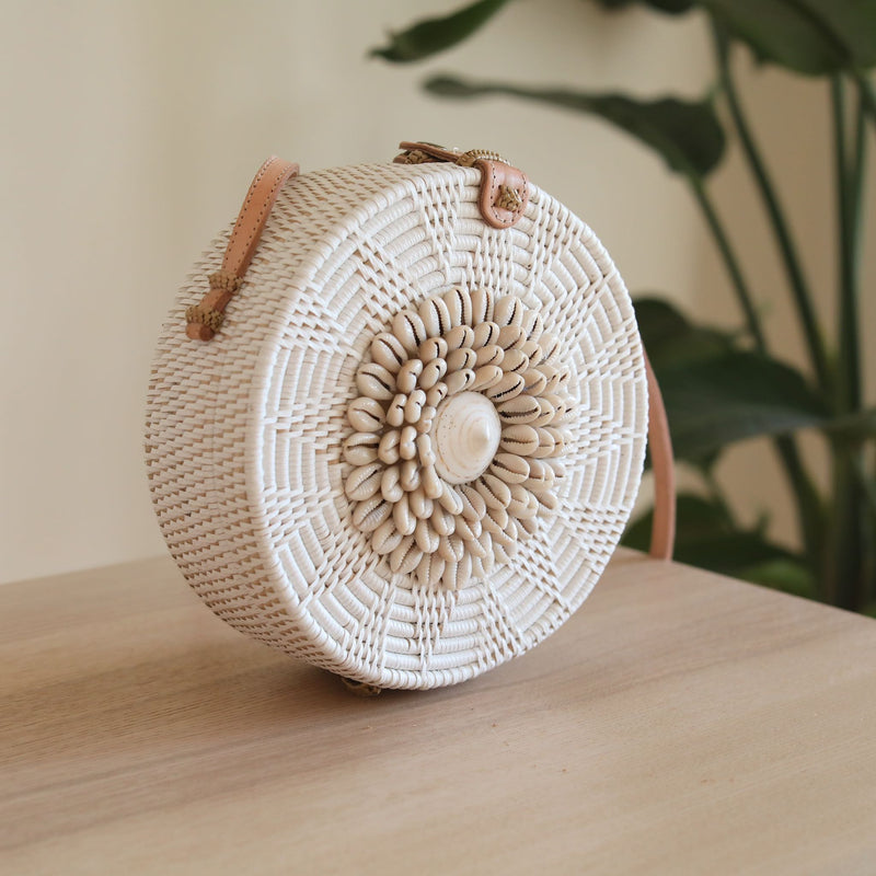A woven rattan crossbody bag, Bali Seashell Rattan Crossbody Bag - Ava, rests on a table with a plant and white wall background. The bag features seashells in its unique design, has a round shape with a handle for carrying over the shoulder, and is made of natural materials like rattan or bamboo.