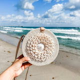 A person holds a round rattan crossbody bag, adorned with real seashells, featuring an adjustable leather strap and batik fabric lining. The background showcases a sandy beach setting under partly cloudy skies.