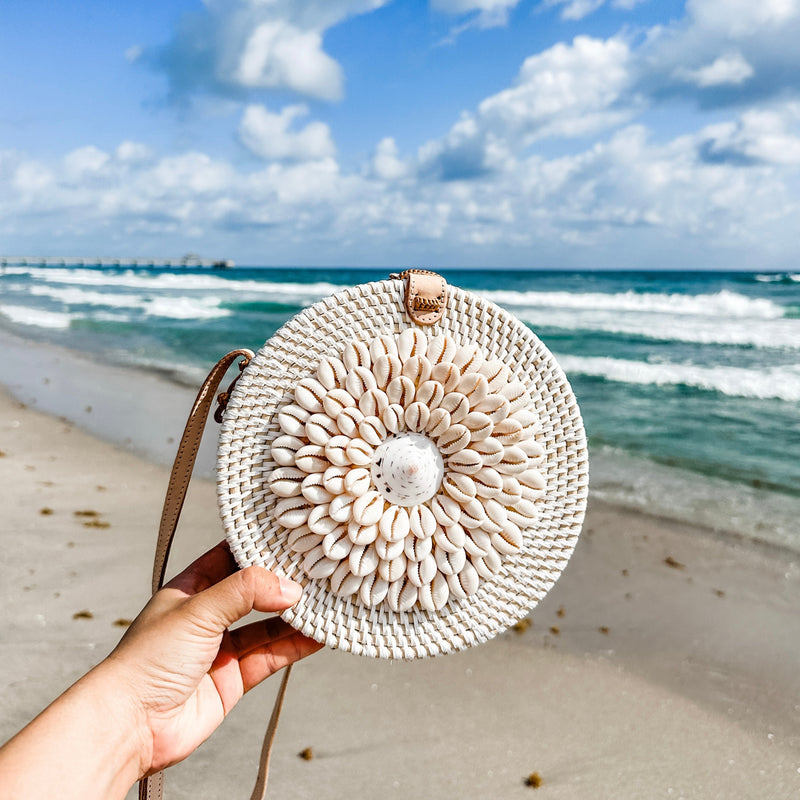 A person holds a round rattan crossbody bag, adorned with real seashells, featuring an adjustable leather strap and batik fabric lining. The background showcases a sandy beach setting under partly cloudy skies.