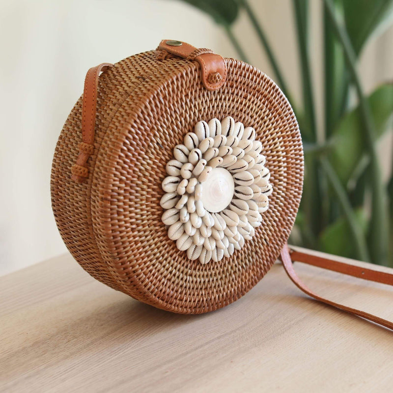 A Bali Round Rattan Crossbody Bag, adorned with real seashells, rests on a wooden table. The bag's natural rattan material is visible, along with its adjustable leather strap and beautiful batik fabric lining.