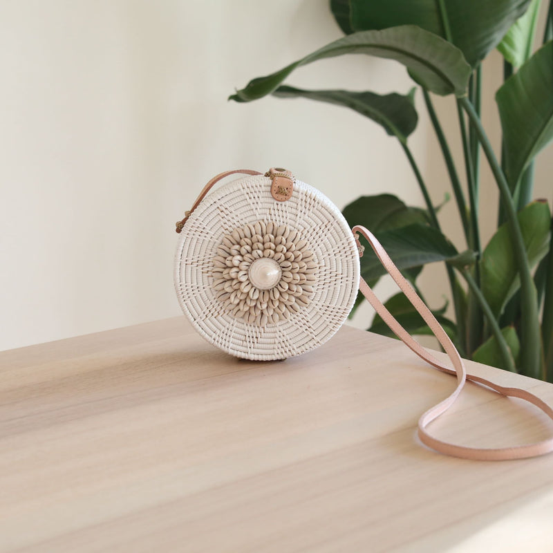 A woven rattan crossbody bag, Bali Seashell Rattan Crossbody Bag - Ava, resting on a table with a plant and white wall in the background. The product features a round top, seashells along its edge, an adjustable leather strap, and is lined with batik fabric.