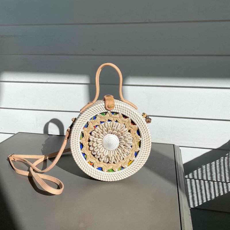 A woven rattan crossbody bag with a seashell design is placed against a white wall, showcasing its natural materials and earthy tones. The round-shaped bag features a handle on top and an adjustable strap for carrying over the shoulder, with shades of brown, cream, and blue dominating the color scheme. Intricate seashell detailing in green, yellow, and white adds a touch of whimsy to this eco-friendly and fair-trade handcrafted piece.