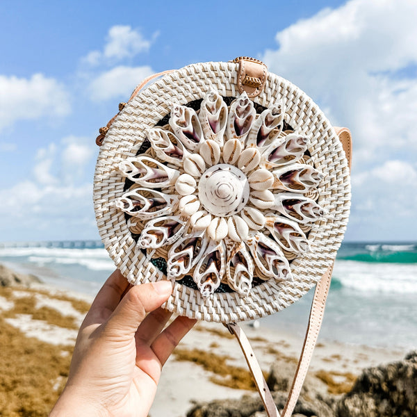 A person holds up a woven crossbody bag adorned with real seashells, featuring a light brown color and circular design on its front. The adjustable leather strap allows for comfortable carrying over the shoulder or across the body. The setting suggests an outdoor lifestyle near a beach, as shown by the sand and seaweed in the background.