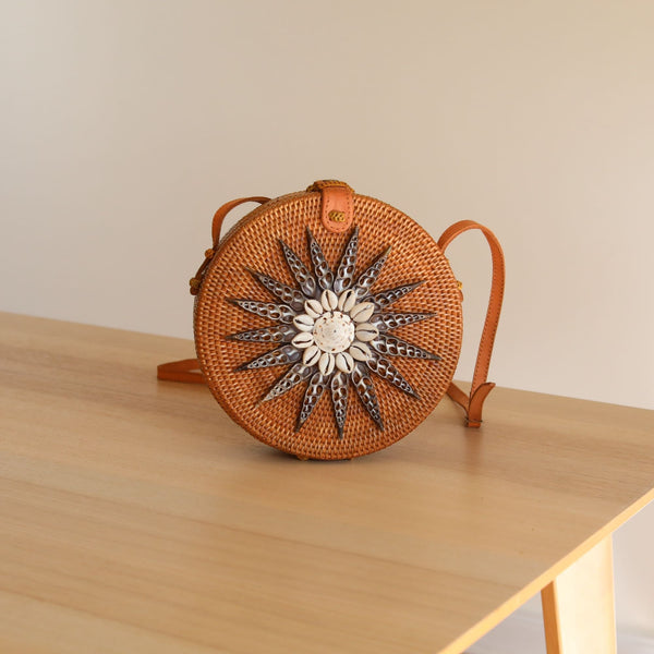 A small, round woven bag with a star-like design on its front rests on a wooden surface. The bag is made from rattan or a similar natural material and features an adjustable strap for crossbody wear. Its batik lining adds a touch of tradition, while the snap closure ensures security.