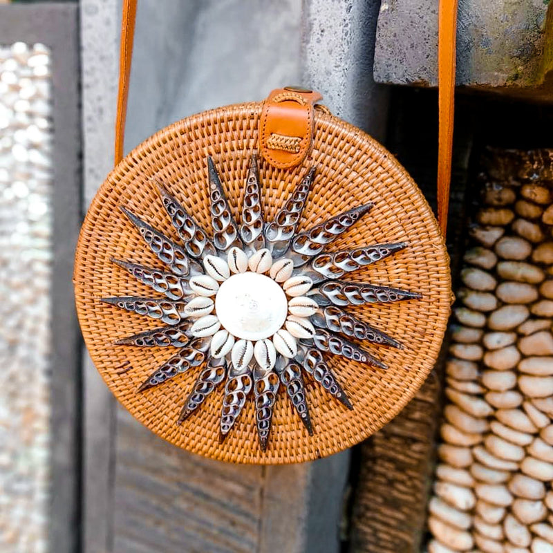 A Bali Seashell Rattan Crossbody Bag - Luna with a sunburst design made up of various seashells in different sizes and colors. The bag is circular, featuring an adjustable strap for carrying over the shoulder, and has a batik lining for durability and traditional aesthetics. A snap closure provides secure fastening. Handmade in Bali from eco-friendly materials, this unique bag showcases coastal elegance with real seashell embellishments and rustic rattan craftsmanship.