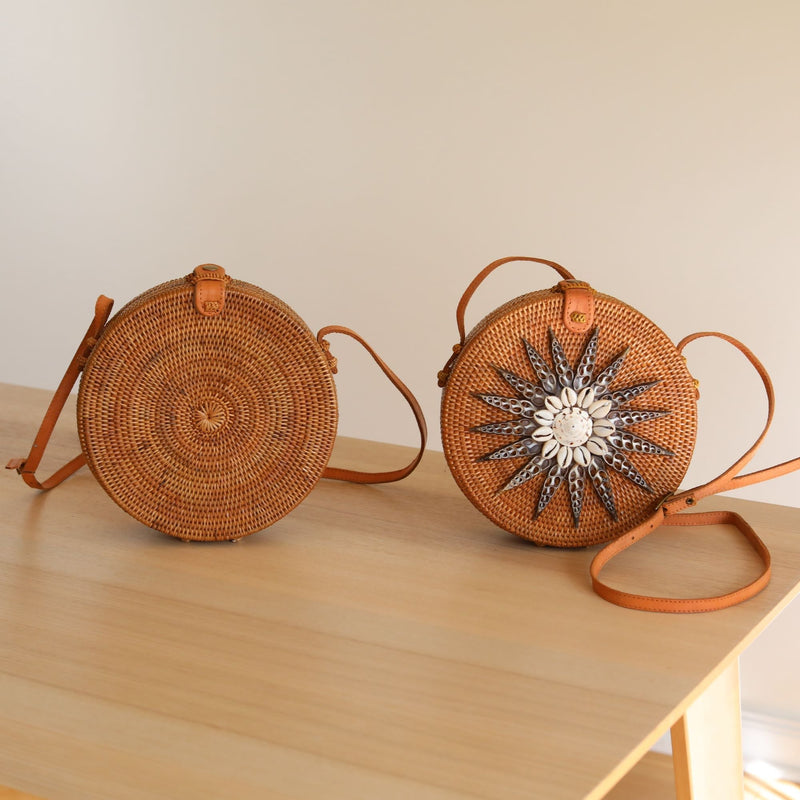 Two woven bags, one larger than the other, rest on a light-colored surface against a plain wall. The front of each bag features a circular design with a star-like pattern within. Brown handles adorned with seashells connect to both bags. The image showcases the intricate details and natural materials used in their construction.