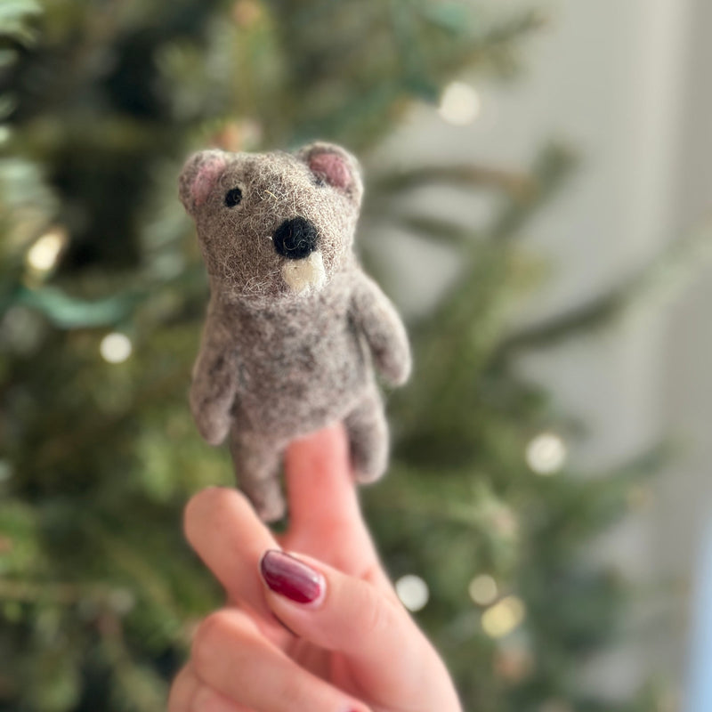 A cozy, handcrafted wool felt beaver finger puppet is held by a hand with red nails, set against a blurred, warmly lit Christmas tree. The charming beaver features a textured grayish-brown body, small black nose, tiny black eyes, and pink ears, highlighting its detailed needle-felted design.
