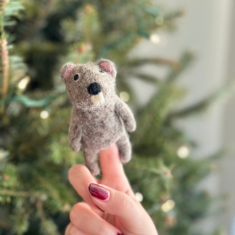 A light brown needle felted beaver finger puppet, crafted from 100% wool, is held by a female hand with dark red nail polish against a softly blurred Christmas tree background. This handmade felt beaver finger puppet features a black nose and small ears, showcasing its delightful 3D design.