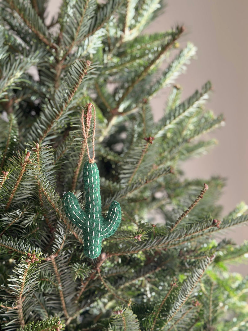 A green Wool Felt Cactus Christmas Ornament hangs delicately from a branch of a lush green Christmas tree. This festive cactus ornament features vertical rows of white stitches mimicking spines and has a hemp string attached for easy hanging, adding a touch of desert charm.