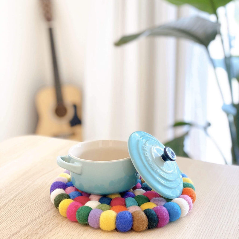 A vibrant, 8-inch diameter circular felt ball pot trivet is displayed on a light wooden table, with a light blue ceramic pot resting on it and its lid nearby. This fair-trade, rainbow-colored wool felt trivet, ethically handmade from hand-stitched organic lamb wool felt balls, showcases red, yellow, white, blue, green, purple, and brown hues. It adds a warm, handcrafted touch to any kitchen or dining space.