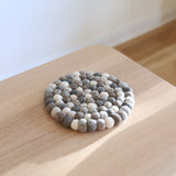 A circular gray felt ball pot trivet, ethically handcrafted from organic wool felt, rests on a light wood surface bathed in soft, natural light. This unique pot trivet, made of tightly arranged felt balls in varying gray and white tones, highlights its tactile texture and the skilled craftsmanship of Nepalese female artisans. It offers understated elegance for home decor.