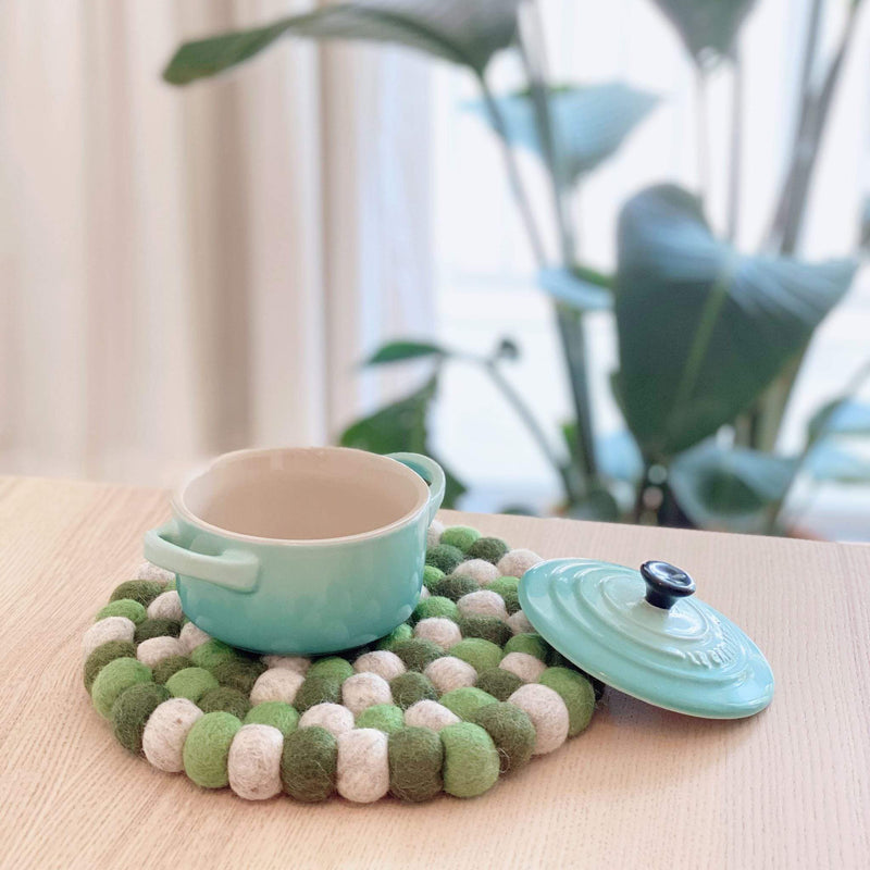 A circular Green Tone felt ball pot trivet is displayed on a light-colored wooden table, with a small blue pot resting beside it. This unique, handcrafted trivet, made from ethically sourced 100% organic lamb wool felt balls, features varying shades of green and white hand-stitched into a textured, heat-resistant design. It brings a calm, natural atmosphere, supporting Nepalese female artisans.