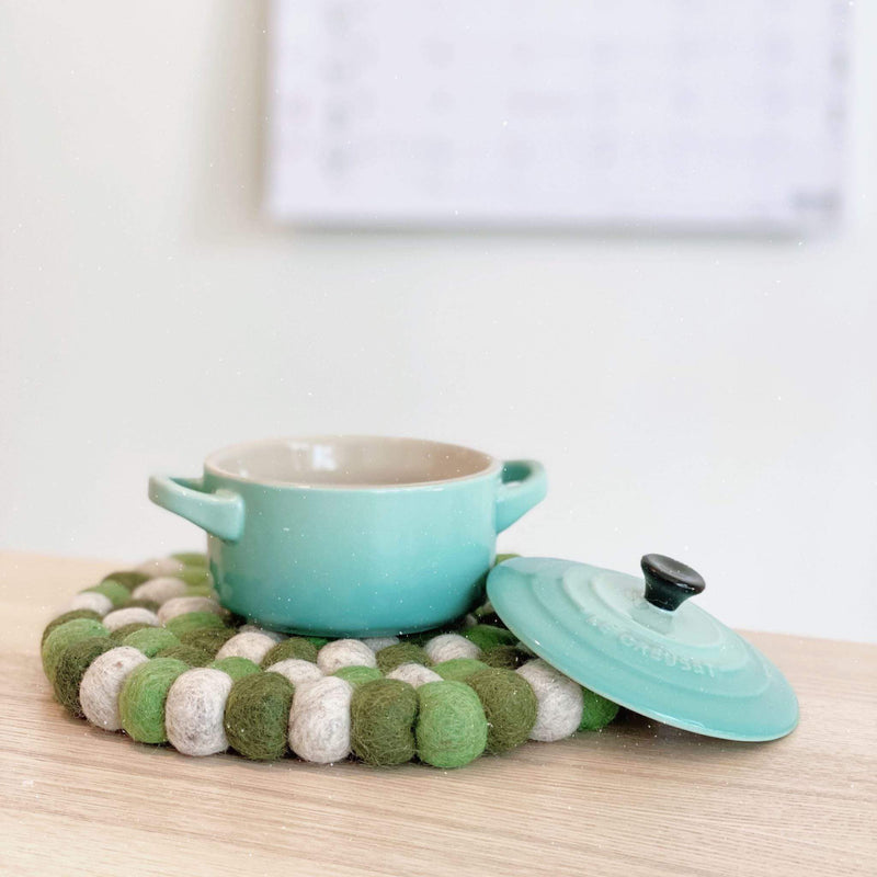 A circular wool felt pot trivet, featuring a blend of green and white organic lamb wool felt balls, is displayed on a wooden surface. A small, light blue pot rests atop this 8-inch handcrafted trivet, highlighting its heat-resistant design. Ethically made by Nepalese female artisans, each felt ball is meticulously hand-stitched.