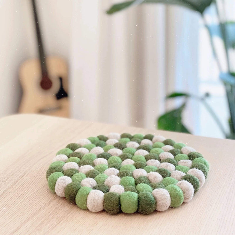 A circular Ø8" green and white felt ball pot trivet, handcrafted from 100% organic lamb wool, rests on a warm wooden table. This ethically handmade wool felt pot trivet features alternating green and white felt balls meticulously stitched together, showcasing its unique craftsmanship. The soft lighting and natural setting with a blurred guitar in the background enhance its inviting, homely feel.