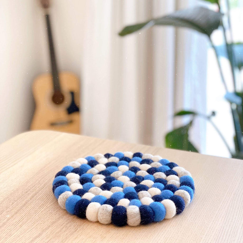 A round, 8-inch Circle Felt Ball Pot Trivet, handcrafted from individual, hand-stitched 100% organic lamb wool felt balls in varying blue and white shades, rests on a light wooden surface. This environmentally friendly and fair-trade piece, ethically made by Nepalese female artisans, offers both heat protection and a cozy decorative touch for home decor. A blurred guitar and plant in the background suggest a warm, inviting setting.