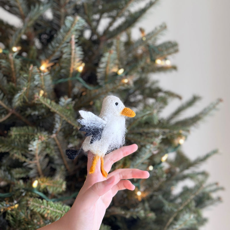 A handcrafted wool felt eagle finger puppet sits on a person's fingers, displayed against the soft, warm glow of a Christmas tree. This unique needle felted eagle puppet is predominantly white with a textured appearance, accented by a bright orange beak and feet, and distinctive black detailing on its wings.