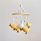 A felt baby mobile, featuring five white call ducks in various poses on a wooden frame, hangs from a ceiling. The mobile's neutral background showcases its playful design, with each duck having distinct expressions and details.