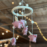 A wool felt baby mobile featuring flying pigs is suspended from the ceiling in a light-filled room, creating a whimsical nursery scene. Several light-pink wool felt pigs, each with small white wings and some adorned with pink bows, hang from white strings. They are suspended from a circular white felt hoop, which is also decorated with colorful felt balls. The design evokes gentle movement and playful imagination.