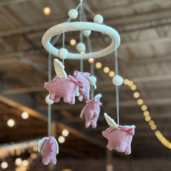 A wool felt baby mobile featuring flying pigs is suspended from the ceiling in a light-filled room, creating a whimsical nursery scene. Several light-pink wool felt pigs, each with small white wings and some adorned with pink bows, hang from white strings. They are suspended from a circular white felt hoop, which is also decorated with colorful felt balls. The design evokes gentle movement and playful imagination.