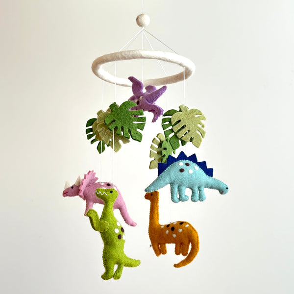 A colorful felt baby mobile, featuring Jurassic dinosaurs and monstera leaves, hangs against a plain white background. A white felt frame suspends a purple Pterosaur, pink Triceratops, green T-Rex with white spots, blue Stegosaurus with dark blue spikes, and an orange Brontosaurus with white spots. The bright felt shapes create a playful prehistoric scene.