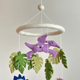A Jurassic Dinosaur felt baby mobile is presented against a plain, light backdrop, creating a soft, playful atmosphere. It features a white felt frame suspending a prominent lavender felt Pterosaur with small, round embellishments and a red beak. Various green felt monstera leaves in different shades hang alongside, all suspended by thin, white strings. This handmade nursery mobile evokes a creative, imaginative prehistoric journey.
