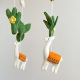 A felt llama baby mobile is shown in a close-up against a soft, neutral gray background. Handcrafted from soft, natural wool felt, the mobile features a central felt hoop from which two white llamas with orange saddles and teal stitching, alongside green cacti with yellow flowers, gently dangle on thin white strings, creating a whimsical and calming atmosphere.
