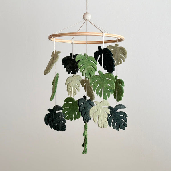 A wooden mobile featuring felt monstera leaves in shades of green, suspended from a white string against a plain background. The leaves are arranged to create a sense of floating or suspension, evoking a calm and serene atmosphere reminiscent of nature.