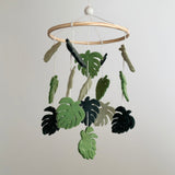 A wooden mobile hangs from a white ceiling, featuring a collection of felt monstera leaves in shades of green. The leaves appear to move due to the string's tension, creating a sense of natural growth and movement. Crafted with wool felt, this baby mobile brings the soothing essence of nature indoors.