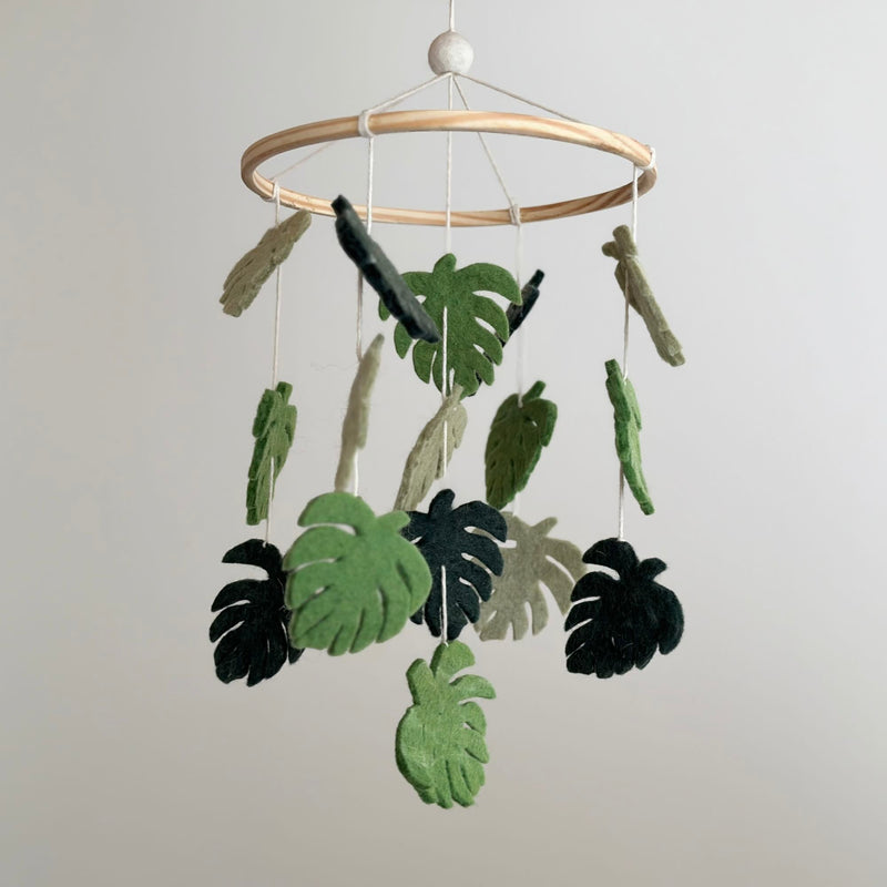 A wooden mobile hangs from a white ceiling, featuring a collection of felt monstera leaves in shades of green. The leaves appear to move due to the string's tension, creating a sense of natural growth and movement. Crafted with wool felt, this baby mobile brings the soothing essence of nature indoors.