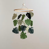 A felt baby mobile, featuring green wool felt leaves in various shapes and sizes, including what appear to be monstera leaves, hangs from a wooden hanger. The mobile's natural design brings the soothing essence of nature indoors, creating a tranquil ambiance perfect for nurseries.