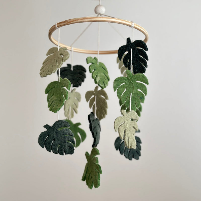 A wooden mobile hangs from a white hook, featuring a collection of felt monstera leaves in various shades of green. The leaves appear to float or suspend in the air, creating a serene ambiance.