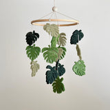 A wooden mobile hangs from a white ceiling, featuring a collection of felt monstera leaves in shades of green. The leaves have darker centers and lighter edges for a realistic appearance, suspended as if floating in the air.
